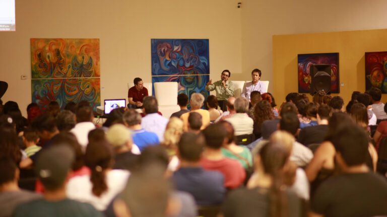 Sala llena durante una charla del festival, con el público de espaldas y los ponentes al frente entre pinturas de gran formato